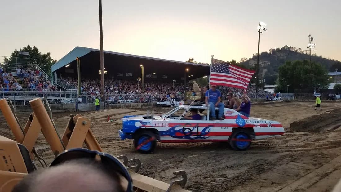 Dirt arena at dusk, a flag-liveried derby car crossing in front of a packed covered grandstand.