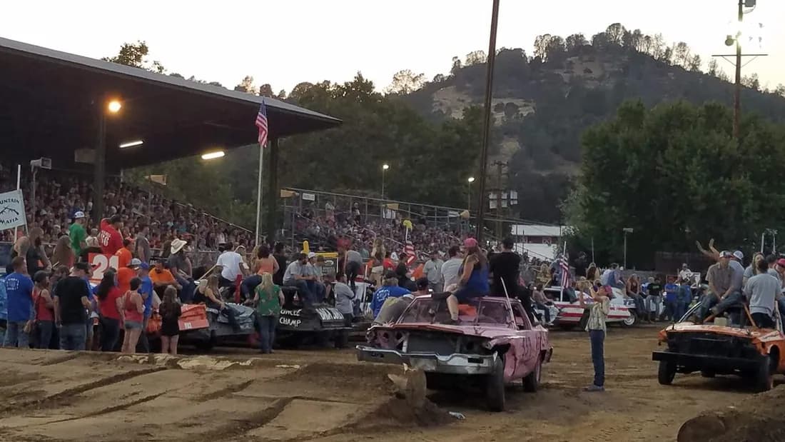 Derby cars staged on the dirt arena with spectators crowding the rail below a full covered grandstand, foothills behind.
